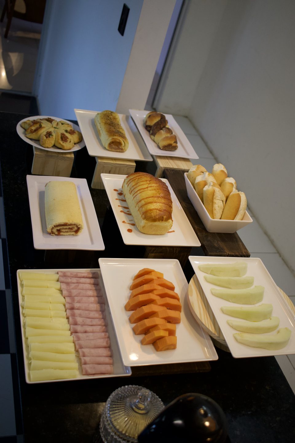 Buffet do café da manhã com bolo de rolo, pães, frios, mamão e melão em travessas