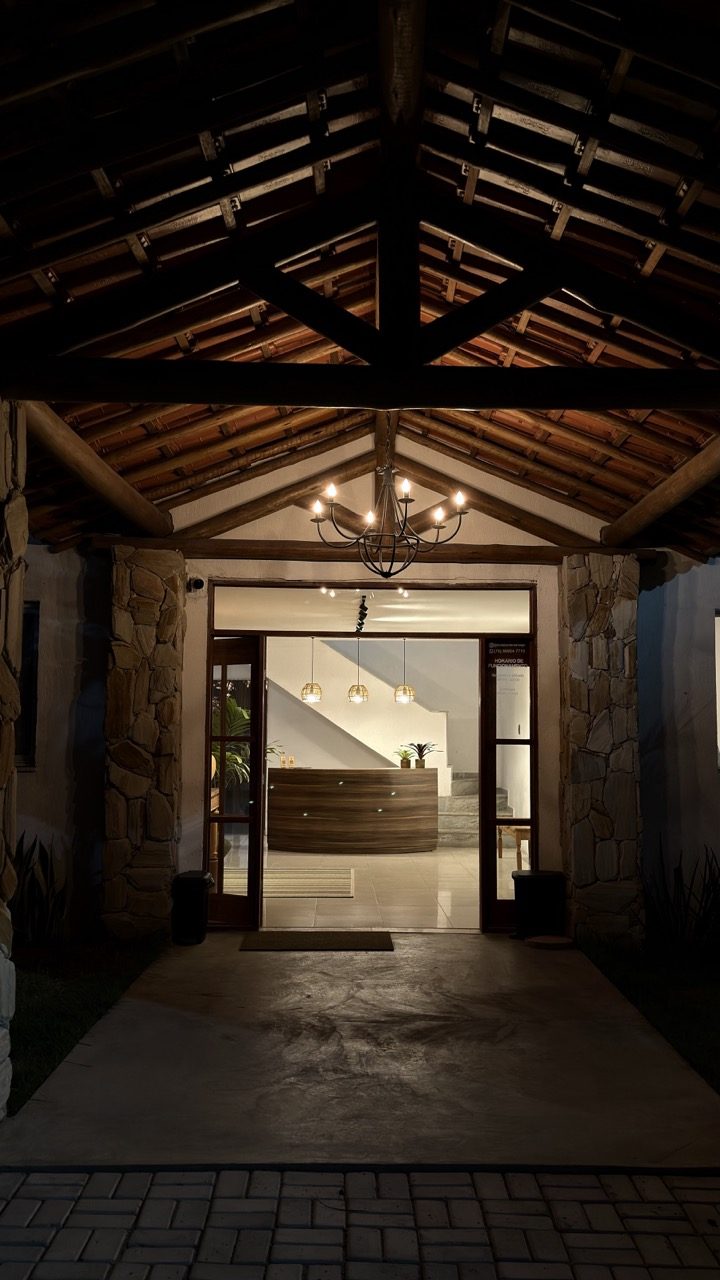 Pórtico de madeira com lustre aceso, com a recepção visível ao fundo pela porta de vidro