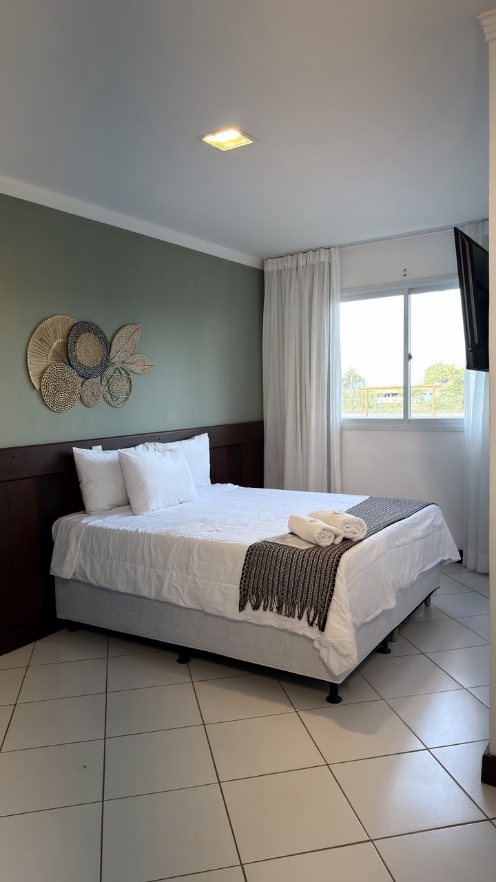 Quarto com cama de casal, parede verde sálvia, cabeceira de madeira e enxoval branco