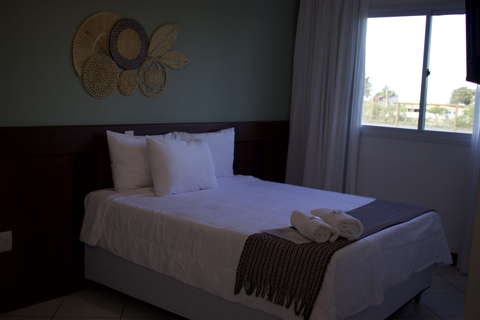 Cama de casal com enxoval branco, parede verde sálvia e peças de fibra trançada na cabeceira
